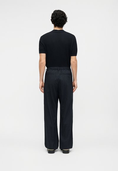 Άνδρας που στέκεται με την πλάτη γυρισμένη, φοράει μαύρο κοντομάνικο πουκάμισο και φαρδιά σκούρα παντελόνια με λεπτές ρίγες, καθώς και μαύρα παπούτσια.