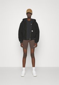 Black zip-up jacket with hood, black t-shirt, brown shorts, and white sneakers. Brown cap. Model stands against a plain background.