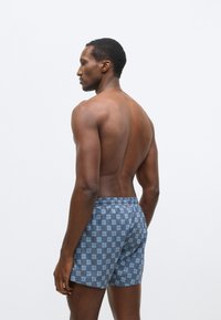 Blauwe boxershorts met een herhalend "HU"-patroon, elastische tailleband en lichtgewicht stof, vanaf de achterkant getoond op een mannelijk model.