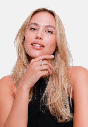 Vrouwelijk model met lang, golvend blond haar, gekleed in een zwarte mouwloze top, die een sprankelende zilveren ring op haar rechterhand toont.