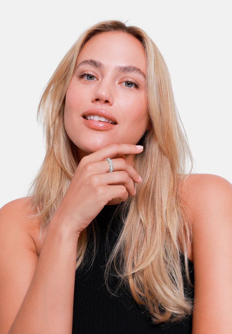 Kvinne modell med langt, bølgete blondt hår, iført en svart ermeløs topp, som viser frem en glitrende sølvring på høyre hånd.