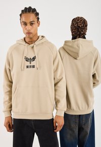 Mand, der står med ansigtet fremad iført en beige hættetrøje med en møl-grafik og japansk tekst, en anden person med leopard-hovedbeklædning står med ryggen til i en beige hættetrøje.