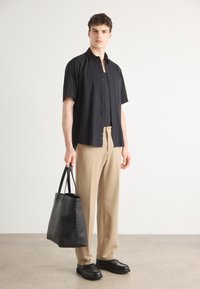 Camicia nera a maniche corte con bottoni, pantaloni beige e borsa a tracolla in pelle nera. Modella che indossa scarpe con plateau nere, in piedi su una superficie neutra.