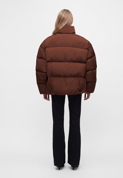 Personne aux longs cheveux blonds portant une doudoune marron et un pantalon noir, debout de dos sur un fond clair uni.