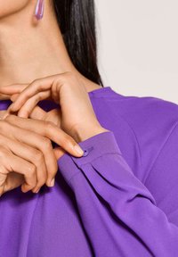 Paarse blouse met lange mouwen. De mouw heeft een knoopsluiting en plooien, met een zachte, lichte textuur. Geaccentueerd door een lila oorbel.
