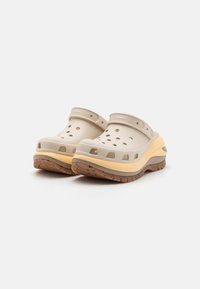 Beige klumper med en myk, teksturert overdel, ventilasjonshull og en strukturert såle med et lagdelt gult og brunt design.