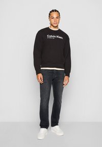 Sweatshirt noir avec logo et texte "Calvin Klein" en blanc, associé à un jean foncé et des baskets blanches. Tissu lisse, design à coupe décontractée.