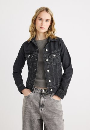 Fiatal nő szőke hajjal, fekete farmerdzsekit, szürke bordázott felsőt és kifakult szürke farmert visel, kezét a zsebében tartva.