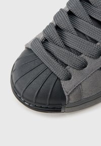 Γκρι αθλητικό παπούτσι με υφή από σουέντ στο πάνω μέρος και μαύρο λαστιχένιο κάλυμμα στα δάχτυλα. Διαθέτει στρογγυλά κορδόνια και λαστιχένια σόλα με οριζόντιες γραμμές.