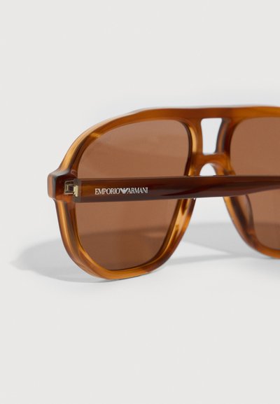 Hnědé oversized sluneční brýle s průhledným hnědým plastovým rámem. Obsahují hnědé tónované čočky a boční branding v bílé barvě na nožičce.