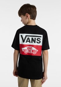 Camiseta de algodón negra con un gran logo blanco de "VANS" y un gráfico rojo de "VANS 'OFF THE WALL'" en la parte posterior. Manga corta, corte relajado.