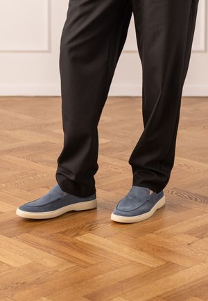 Persona che indossa pantaloni neri e scarpe senza lacci in camoscio blu, in piedi su un pavimento in parquet di legno all'interno.