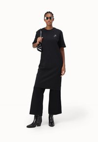 Svart oversized t-skjortekjole med korte ermer, kombinert med vide svarte bukser. Tilbehør inkluderer liten svart håndveske og solbriller.