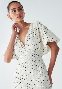 White dress with black polka dots, deep V-neck, short puff sleeves, and a fitted waist. Made of lightweight fabric with a textured finish.