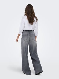 Mujer con cabello largo y ondulado, vistiendo una camiseta blanca de manga larga y vaqueros anchos de color gris desgastado, de pie de espaldas sobre un fondo liso.