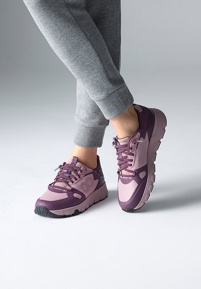 Des chaussures de sport violet et rose avec une tige en maille, une semelle rembourrée et des accents texturés, portées avec un pantalon de survêtement gris.