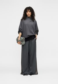 Γκρι φαρδιά μπλούζα με ψηλό λαιμό, σε συνδυασμό με άνετο γκρι παντελόνι φαρδύ στα πόδια. Το μοντέλο κρατά τσάντα με σχέδιο και στριφτό μοτίβο.