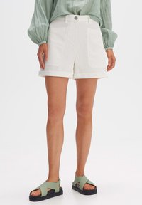 Pantaloni corti bianchi in cotone con orlo risvoltato, tasche frontali e chiusura con bottone in metallo, abbinati a sandali verdi con cinturini su uno sfondo chiaro.