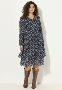 Vestido floral azul marino con mangas largas transparentes, dobladillo de volantes en capas y cintura elástica. Combinado con botas de vaquero marrones.