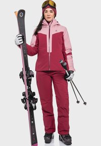 Ski outfit in burgundy and pink, featuring a waterproof jacket with a zipper, matching pants, and black skis with pink accents and poles.