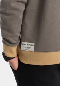 Grå sweatshirt med en gylden ribkant, der bærer en label med teksten "OMBRE MENSWEAR." Glat tekstur, afslappet design og lange ærmer.