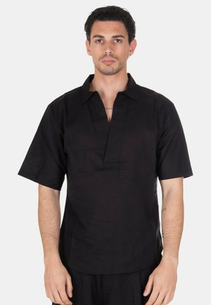 Uomo con capelli corti e scuri che indossa una camicia nera larga a maniche corte con scollo a V e in piedi davanti a uno sfondo chiaro e uniforme.