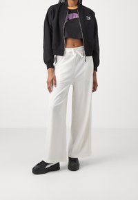 Chaqueta bomber negra, camiseta negra corta con logo morado, pantalones blancos acampanados de canalé y zapatillas negras con detalles blancos.