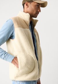 Fleece vest in cream with a smooth tan upper panel, zip pockets, and high collar. Worn over a denim shirt, featuring a relaxed fit.