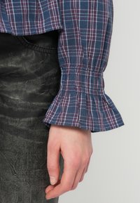 Kék és burgundi kockás ing fodros mandzsettával, textúrált szövet részletekkel, fekete bőr szoknyával párosítva.