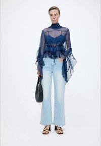 Γυναίκα που φοράει διάφανη μπλε μπλούζα με βολάν, ανοιχτό μπλε τζιν με φαρδύ πόδι, μαύρα σανδάλια με τακούνι και κρατάει μαύρη τσάντα, στέκεται μπροστά σε λευκό φόντο.