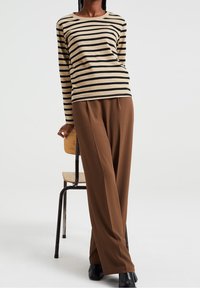 Osoba se opírá o dřevěnou židli, má na sobě dlouhý rukáv tričko s béžovými a černými pruhy, hnědé široké kalhoty a černé boty.