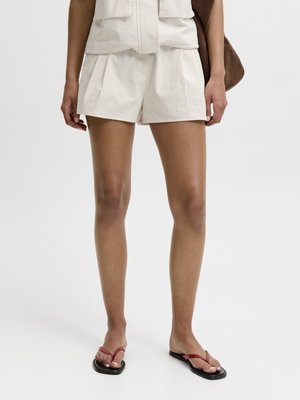 Person iført beige plisserede shorts, en matchende top og sorte flip-flops med tynde røde remme, stående mod en ensfarvet baggrund.