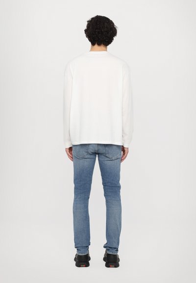 Wit lange mouwen shirt met een relaxte pasvorm, gecombineerd met blauwe skinny jeans en zwarte sneakers. Het shirt heeft een ronde hals.