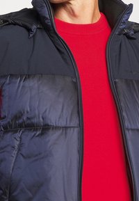 Chaqueta acolchada marina con cierre de cremallera, cuello alto y textura brillante, llevada sobre un suéter texturizado de un rojo brillante. Presenta acentos en los hombros.