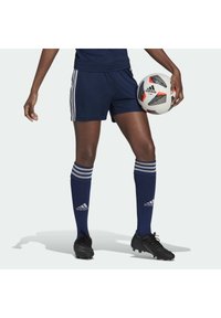 Laivastonsiniset jalkapalloshortsit, joissa on kolme valkoista raitaa, yhdistettynä polven korkeisiin laivastonsinisiin sukkihin, joissa on valkoisia raitoja. Kädessä pidetään mustaa jalkapalloa.