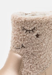 Botine bej căptușite cu blană, cu o textură ondulată. Accente din piele și detalii brodate, inclusiv ochi și stele, pe partea superioară moale.