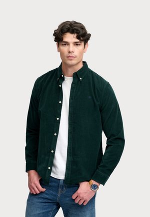 Joven con cabello rizado oscuro que lleva una camisa verde oscuro abotonada sobre una camiseta blanca y vaqueros azules, con un reloj en la muñeca izquierda.