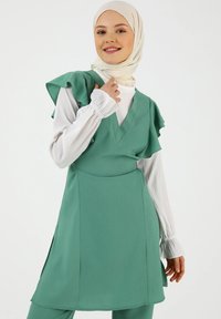 Vihreä kerroksittain oleva mekko, jossa on volangihihat, V-pääntie ja sivuhalkiot; päällä valkoinen pitkähiha-aloitus, jossa on rypytetyt hihansuut. Sujuva tekstuuri.