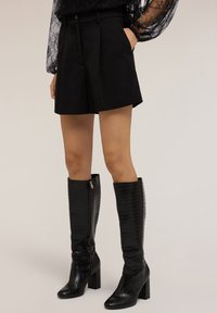 Pantaloni shorts neri su misura con pieghe, realizzati in tessuto liscio, abbinati a stivali neri al ginocchio con embossing in croco, dotati di tacco a blocco e cerniera.