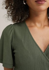 Olivgrön strukturerad blus med korta puffärmar och V-ringning, prydd med vertikala ribbor för extra detalj.
