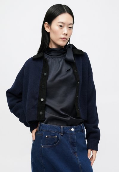 Γυναίκα με ναυτικό μπουφάν με μαύρη επένδυση, μαύρο σατέν πουκάμισο με φιόγκο στον λαιμό και ψηλόμεση μπλε τζιν φούστα, στέκεται μπροστά σε ένα απλό φόντο.