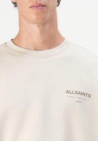 Sweatshirt beige clair avec un col rond et des poignets côtelés. Présente un logo simple en gris foncé sur le devant, affichant le nom de la marque.