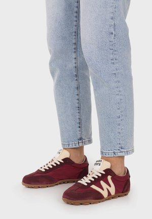 Persona con vaqueros azul claro y zapatillas burdeos con detalles beige, de pie sobre fondo blanco.