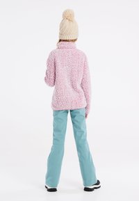 Pull en polaire à motifs rose et blanc, avec un col montant. Associé à un pantalon imperméable bleu clair et des bottes noires. Bonnet en tricot beige avec pompon.