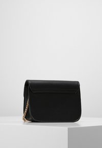 Černá kožená crossbody taška se zlatým řetízkovým popruhem leží na bílém podstavci proti jednobarevnému šedému pozadí.