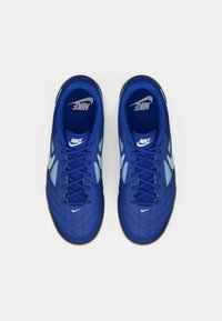 Par nogometnih čevljev Nike v modri barvi za notranje utrjene površine z belimi logotipi swoosh, črnimi vezalkami in podplati iz gume, gledano od zgoraj na belem ozadju.