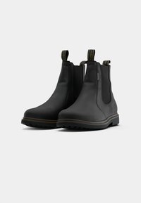 Um par de botins Chelsea pretos em couro com solas castanhas, painéis elásticos laterais e alças de puxar com detalhes da marca.