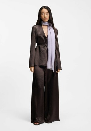 Nainen, joka käyttää tummanruskeita satiinisia leveälahkeisia housuja ja vastaavaa bleiseriä vaalean violetin huivin kanssa, seisoo yksivärisen valkoisen taustan edessä.