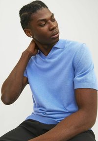 Ljusblå kortärmad v-neck tröja med en slät textur och ribbade ärmslut, kombinerad med mörka byxor. Modellen sitter ner och tittar åt sidan.