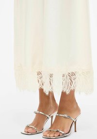 Kremfarget kjole til ankler med intrikate blonder ved kanten; kombinert med sølvmetalliske høye hæler.